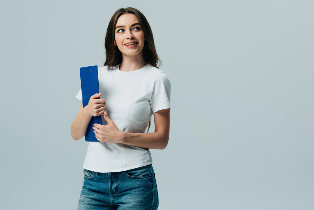 happy beautiful girl in white t-shirt holding clipboard looking away isolated on grey - Photo, Image