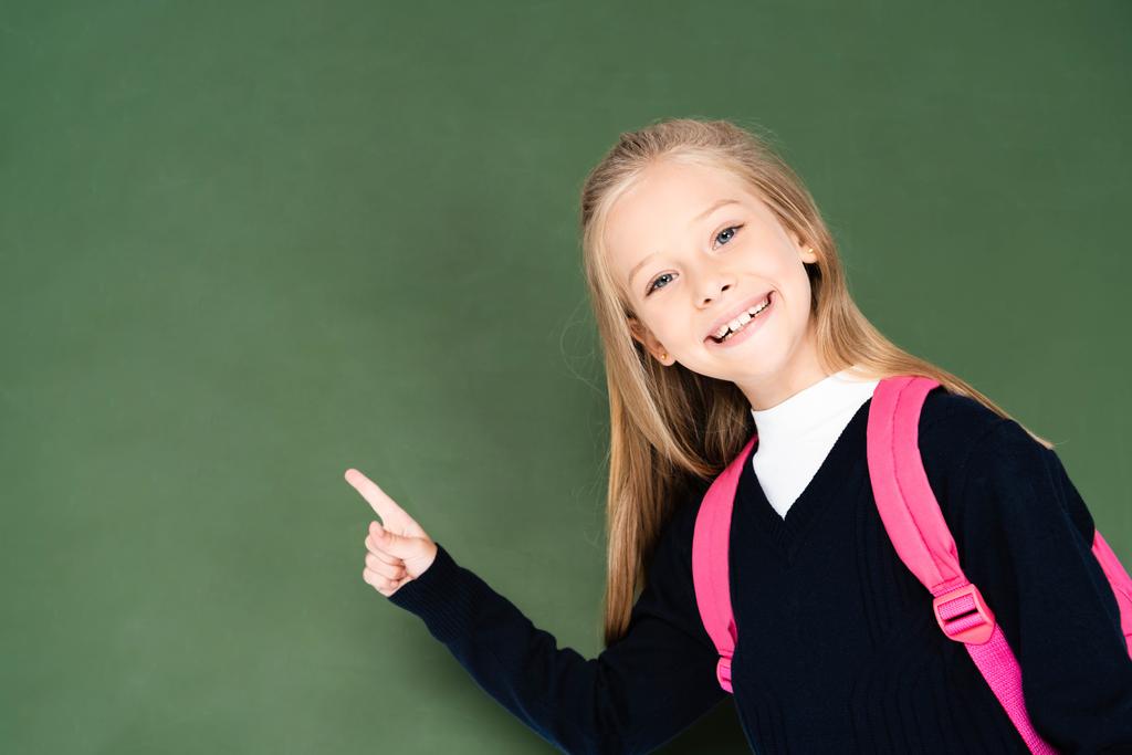 happy schoolgirl pointing with finger at green chalkboard while smiling at camera - Φωτογραφία, εικόνα