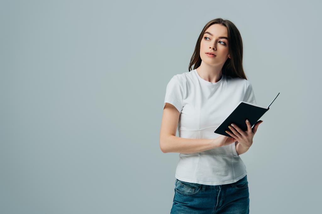 dreamy beautiful girl in white t-shirt holding notebook isolated on grey - Photo, Image