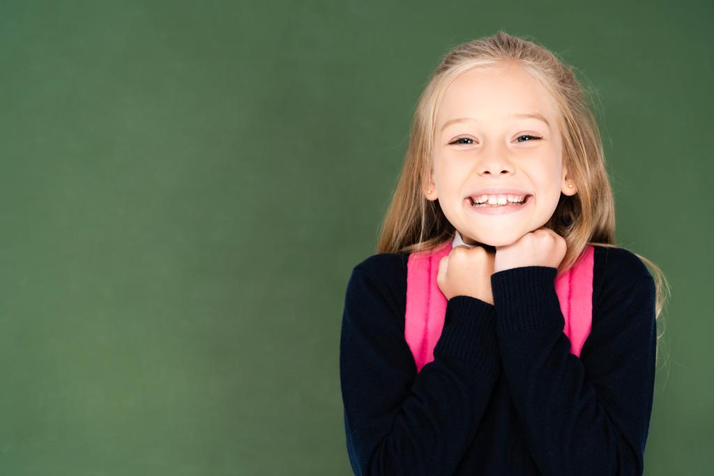 cute, happy schoolgirl smiling at camera while standing near green chalk board - Photo, Image