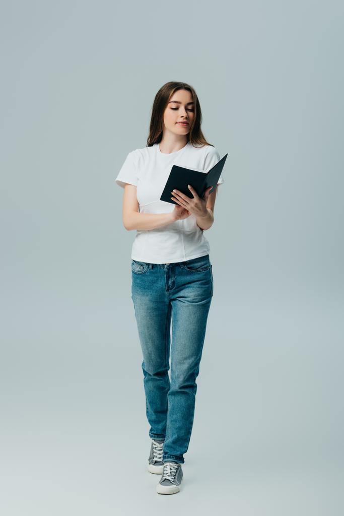 красивая девушка в белой футболке и джинсах, читающая книгу, изолированную на сером
 - Фото, изображение