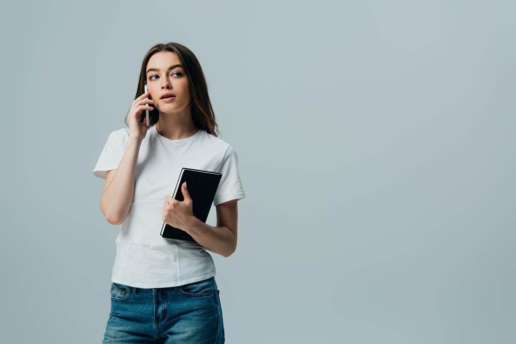 chica hermosa de ensueño en camiseta blanca hablando en el teléfono inteligente y la celebración de portátil aislado en gris
 - Foto, Imagen