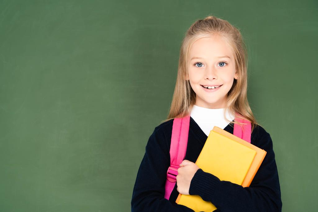 cheerful schoolgirl holding book and smiling at camera while standing near green chalkboard - Photo, Image