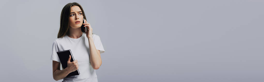 trauriges schönes Mädchen in weißem T-Shirt, das auf dem Smartphone spricht und sein Notizbuch isoliert auf grauem, panoramischem Foto hält - Foto, Bild