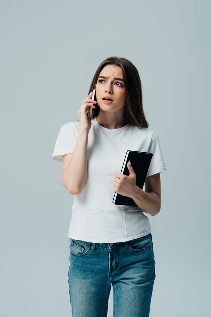 Schockiertes schönes Mädchen in weißem T-Shirt, das auf dem Smartphone spricht und sein Notizbuch isoliert auf grau hält - Foto, Bild