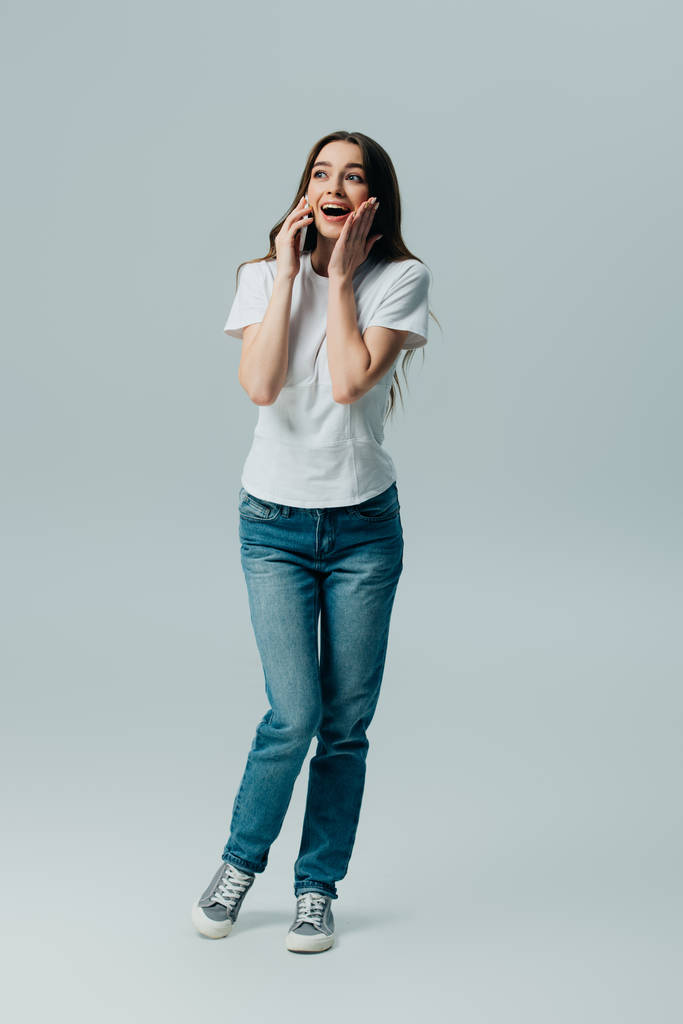 взволнованная, удивленная красивая девушка в белой футболке и джинсах разговаривает на смартфоне изолированы на серый
 - Фото, изображение