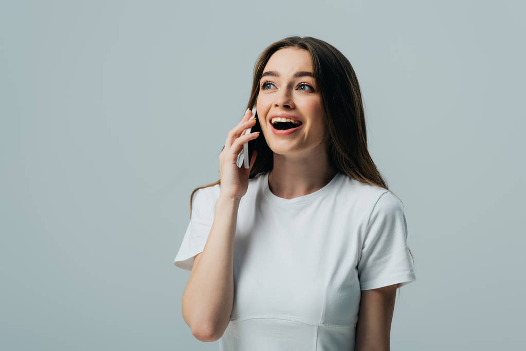 emocionado sorprendido hermosa chica en camiseta blanca hablando en el teléfono inteligente aislado en gris
 - Foto, Imagen
