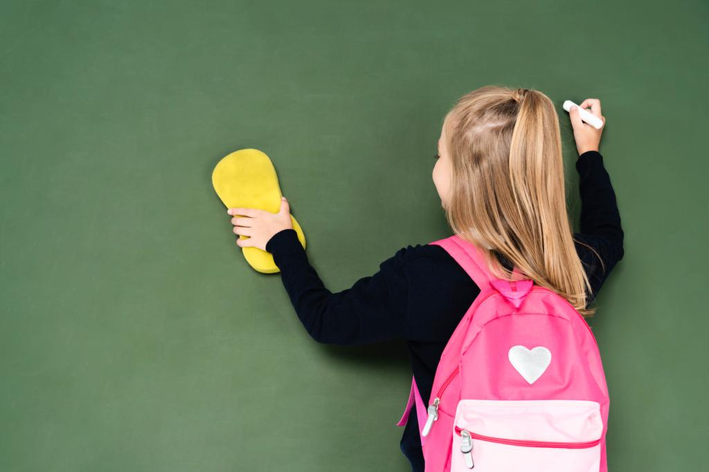 back view of schoolgirl wiping chalkboard with sponge while holding piece of chalk  - Photo, Image