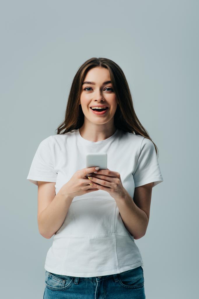 ενθουσιασμένος όμορφο κορίτσι με λευκό t-shirt χρησιμοποιώντας smartphone απομονωθεί σε γκρι - Φωτογραφία, εικόνα