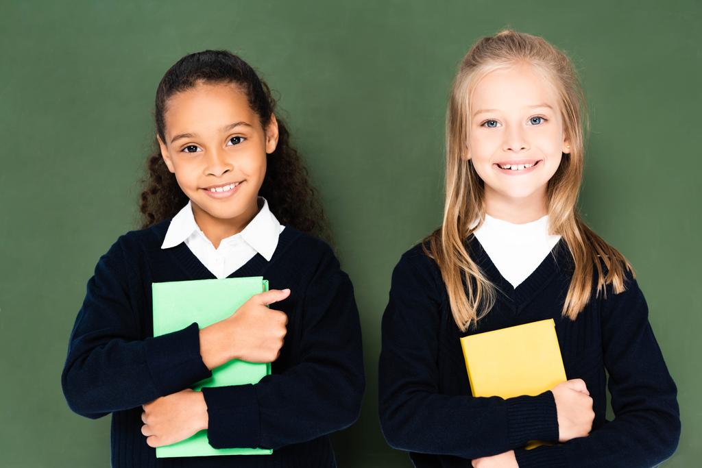 две милые мультикультурные школьницы улыбаются в камеру, стоя рядом с доской и держа книги
 - Фото, изображение