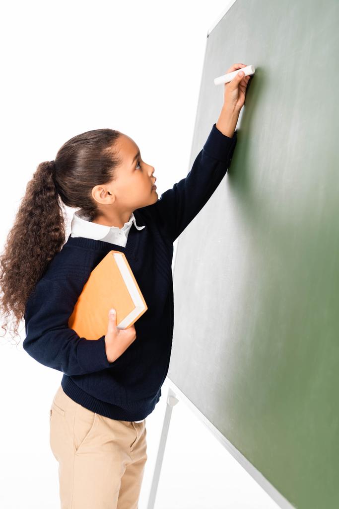aranyos afro-amerikai iskoláslány írásban Chalkboard miközben könyvet elszigetelt fehér - Fotó, kép