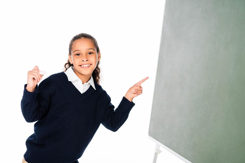 vrolijke African American schoolmeisje wijzend met vinger op groene schoolbord geïsoleerd op wit - Foto, afbeelding