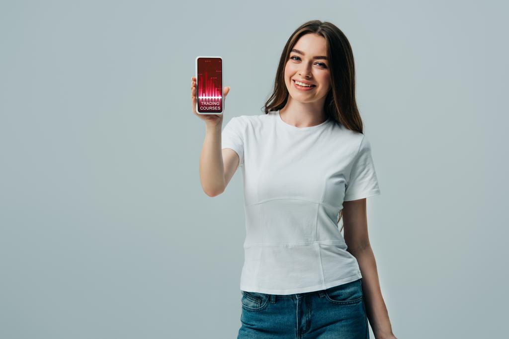 feliz hermosa chica en camiseta blanca que muestra el teléfono inteligente con cursos de comercio aplicación aislada en gris
 - Foto, Imagen