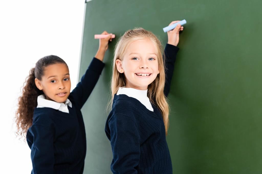 twee glimlachende multiculturele schoolmeisjes schrijven op schoolbord terwijl ze naar de camera kijken  - Foto, afbeelding