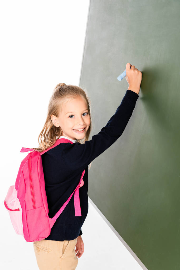 cheerful schoolgirl writing on green chalkboard while smiling at camera - Photo, Image