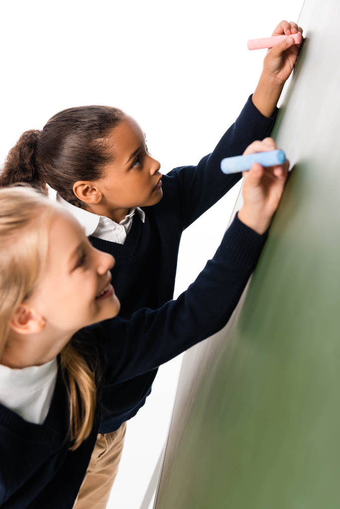two adorable multicultural schoolgirls writing on green chalkboard - Photo, Image