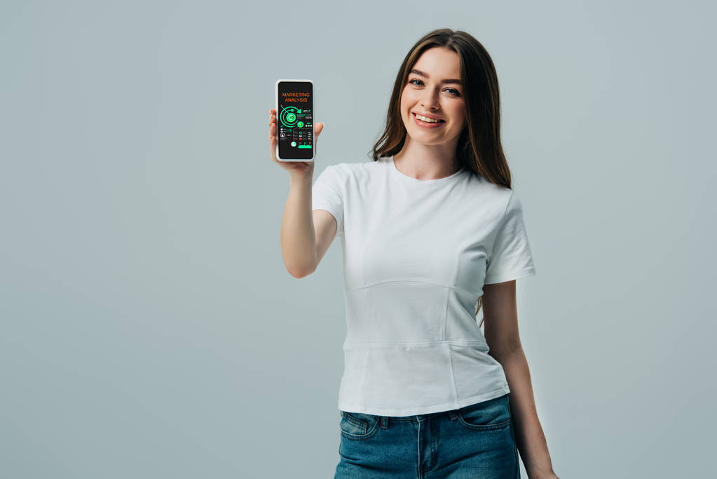 szczęśliwy piękna dziewczyna w białym t-shirt przedstawiający smartfon z aplikacją do analizy marketingowej na szarym tle - Zdjęcie, obraz