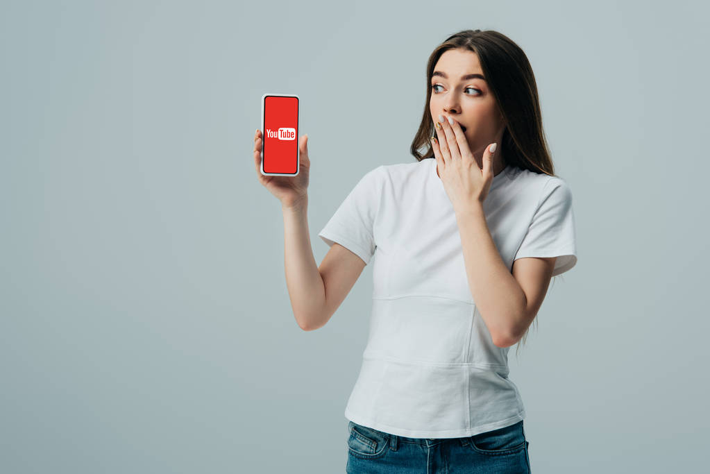 KYIV, UCRANIA - 6 de junio de 2019: impactada hermosa chica en camiseta blanca que muestra el teléfono inteligente con la aplicación de youtube aislada en gris
 - Foto, imagen