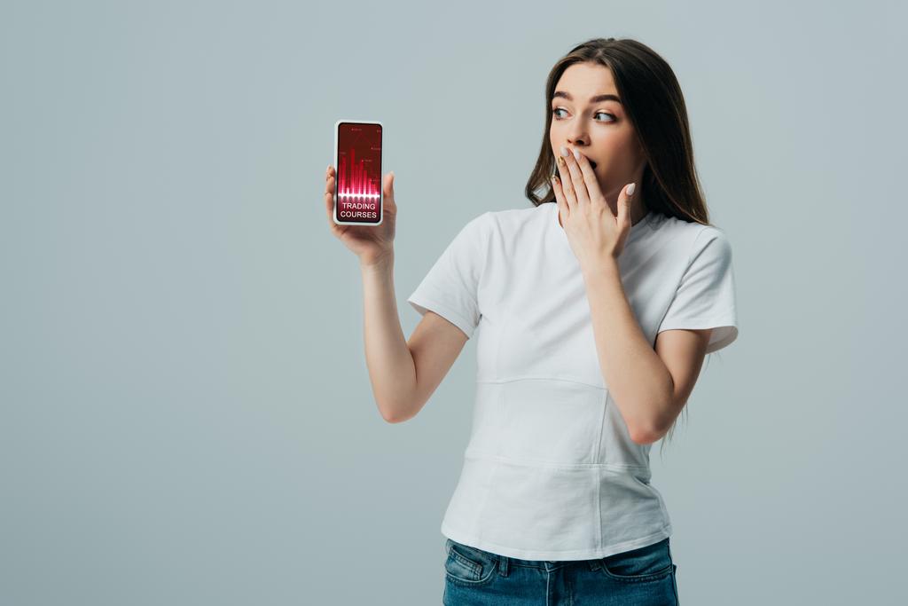 šokovaná krásná dívka v bílém tričku, ukazující smartphone s obchodními kurzy aplikace izolované na šedé - Fotografie, Obrázek