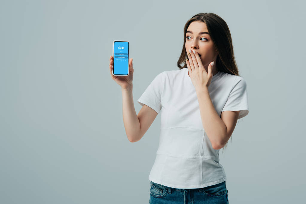 KYIV, UKRAINE - kesäkuu 6, 2019: järkyttynyt kaunis tyttö valkoisessa t-paidassa, joka näyttää älypuhelimen Skype-sovelluksella harmaalla eristettynä
 - Valokuva, kuva
