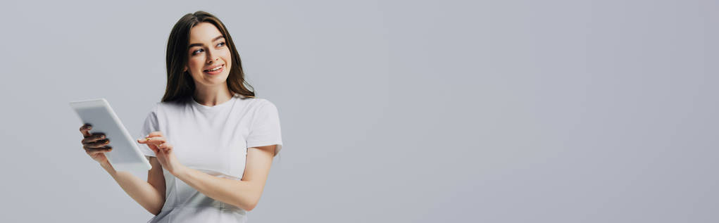 menina feliz em t-shirt branca segurando tablet digital e olhando para longe isolado em cinza, tiro panorâmico
 - Foto, Imagem
