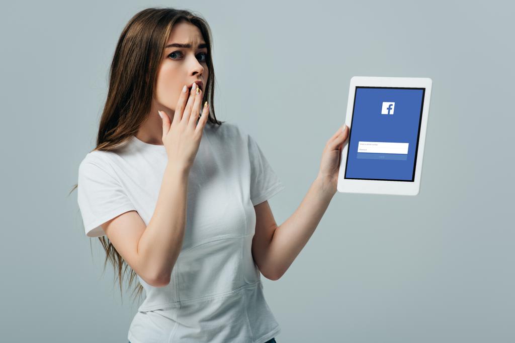 Kijev, Ukrajna-június 6, 2019: megdöbbentett szép lány, fehér póló mutatja a digitális tabletta Facebook App izolált szürke - Fotó, kép