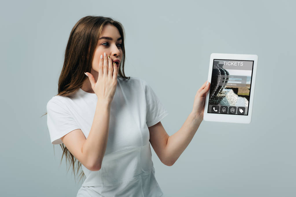 потрясена красивая девушка в белой футболке показывая цифровой планшет с билетами приложение изолированы на серый
 - Фото, изображение