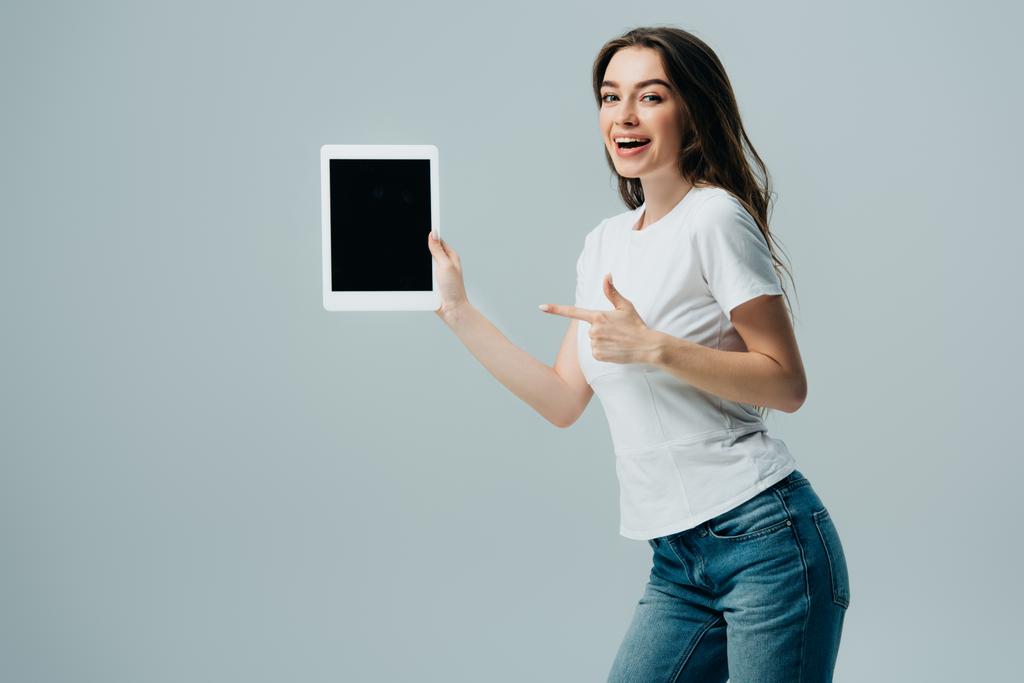 glimlachend mooi meisje in wit t-shirt wijzend met de vinger op digitale tablet met blanco scherm geïsoleerd op grijs - Foto, afbeelding