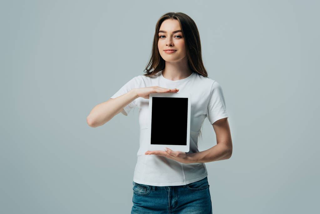 lächelndes schönes Mädchen in weißem T-Shirt, das ein digitales Tablet mit leerem Bildschirm zeigt, isoliert auf grau - Foto, Bild