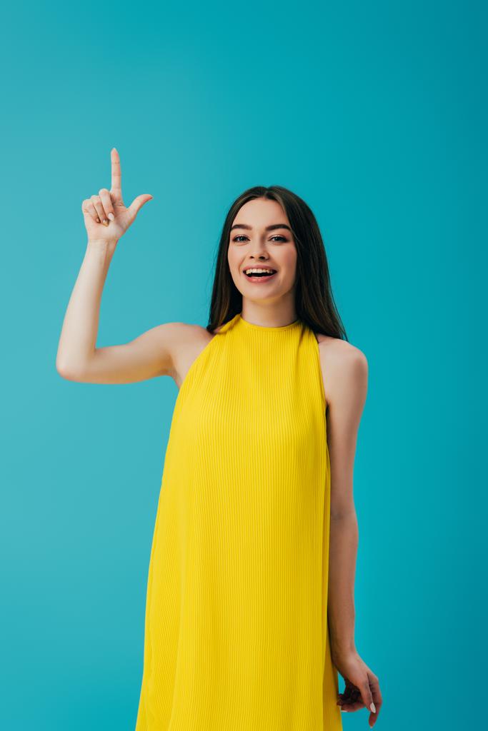 feliz hermosa chica en vestido amarillo apuntando con el dedo hacia arriba aislado en turquesa
 - Foto, Imagen