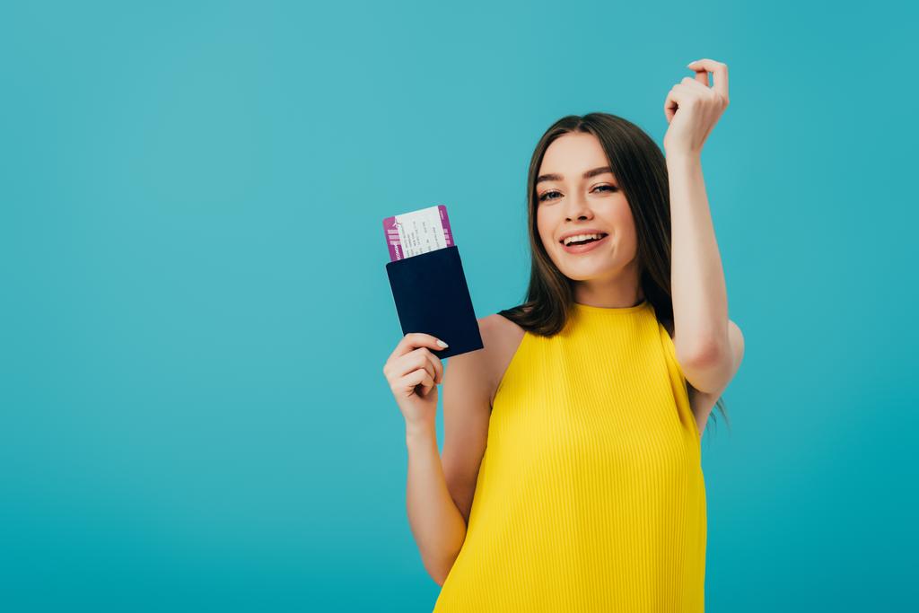 веселая брюнетка в желтом платье танцует с паспортом с авиабилетом изолированы на бирюзовый
 - Фото, изображение