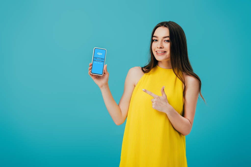 КИЕВ, УКРАИНА - 6 ИЮНЯ 2019 года: счастливая красивая девушка в жёлтом платье, указывающая пальцем на смартфон с приложением Skype, изолированным на бирюзовом
 - Фото, изображение