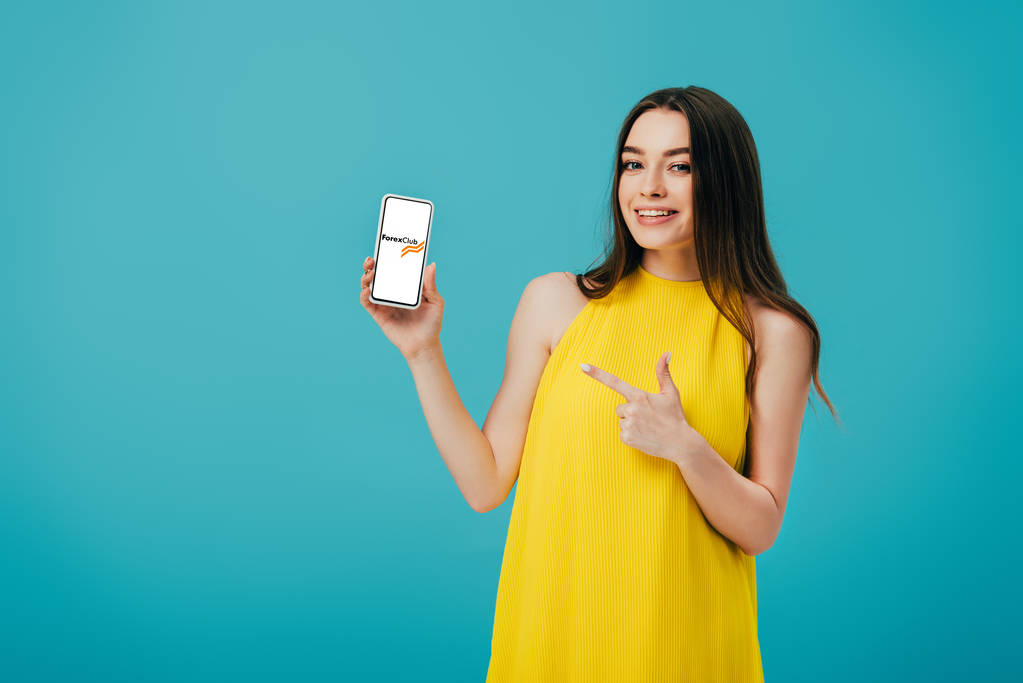 KYIV, UKRAINE - JUNHO 6, 2019: menina bonita feliz em vestido amarelo apontando com o dedo para o smartphone com aplicativo clube forex isolado em turquesa
 - Foto, Imagem