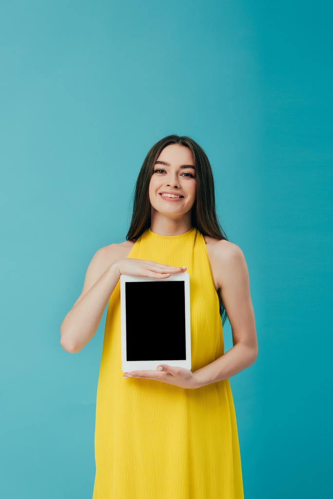 улыбающаяся брюнетка в желтом платье показывает цифровой планшет с чистым экраном, изолированным на бирюзовом
 - Фото, изображение