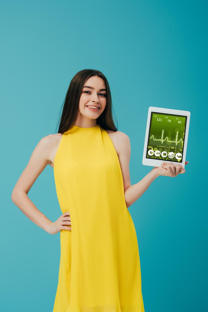 chica morena sonriente en vestido amarillo mostrando tableta digital con aplicación sanitaria aislada en turquesa
 - Foto, imagen