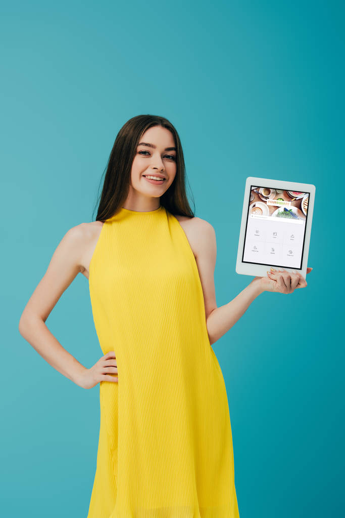 KYIV, UCRANIA - 6 DE JUNIO DE 2019: niña sonriente en vestido amarillo con la mano en la cadera mostrando tableta digital con aplicación cuadrada aislada en turquesa
 - Foto, imagen