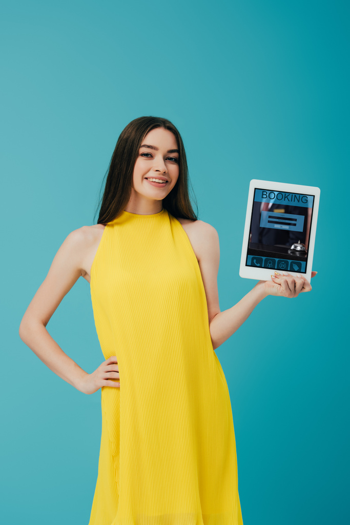 Gelukkig brunette meisje in gele jurk tonen digitale tablet met Booking App geïsoleerd op Turquoise - Foto, afbeelding