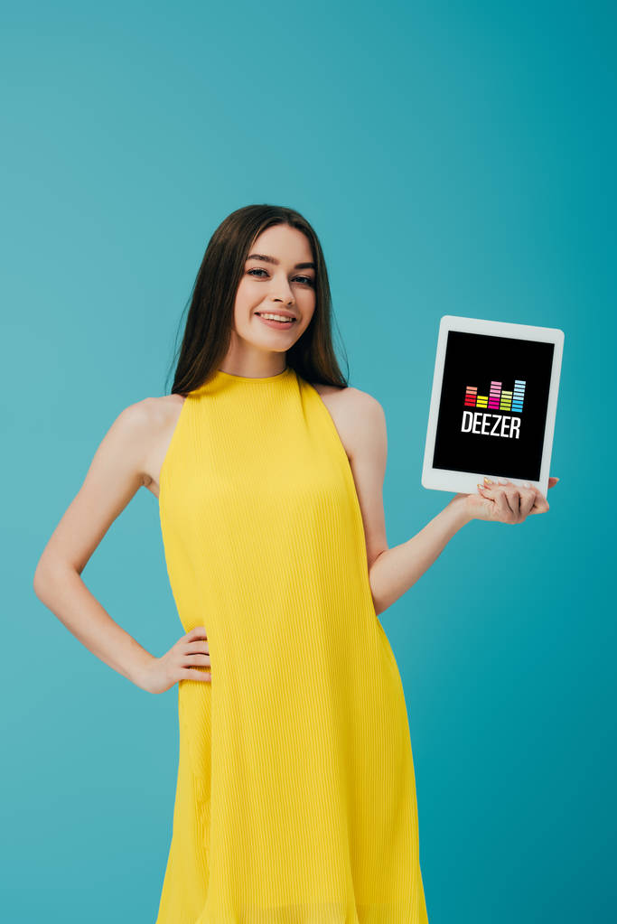 КИЕВ, УКРАИНА - 6 ИЮНЯ 2019 года: улыбающаяся девушка в желтом платье с рукой на бедре показывает цифровой планшет с приложением Deezer изолированы на бирюзовом
 - Фото, изображение