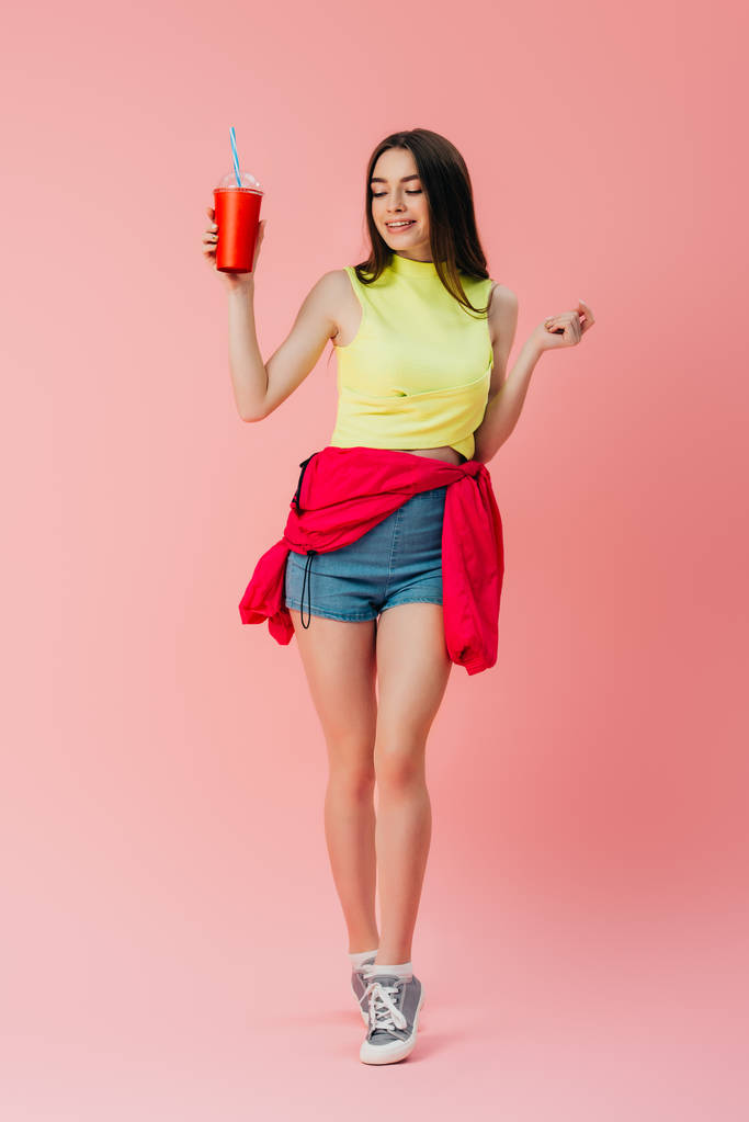 Вид в полный рост красивой счастливой девушки в яркой одежде, держащей газировку в пластиковой чашке, изолированной на розовом фоне
 - Фото, изображение