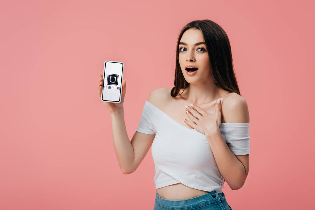 Kyjev, Ukrajina-6. června 2019: krásná šokovaná dívka držící smartphone s superspartánské aplikací izolované na růžovém - Fotografie, Obrázek