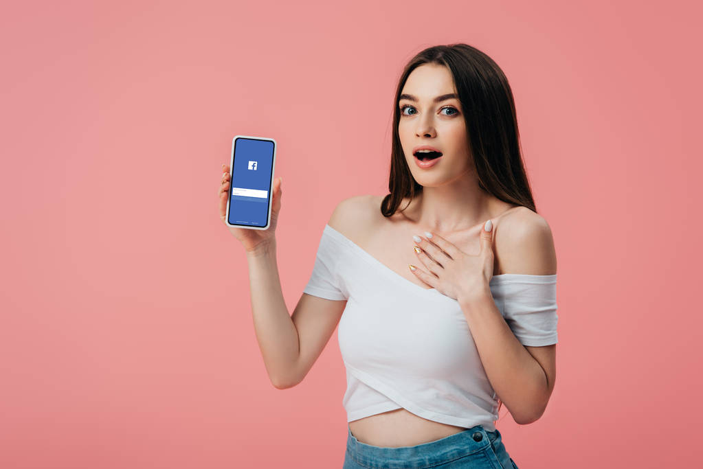 Kiev, Oekraïne-6 juni 2019: mooi geschokt meisje houden smartphone met Facebook app geïsoleerd op roze - Foto, afbeelding