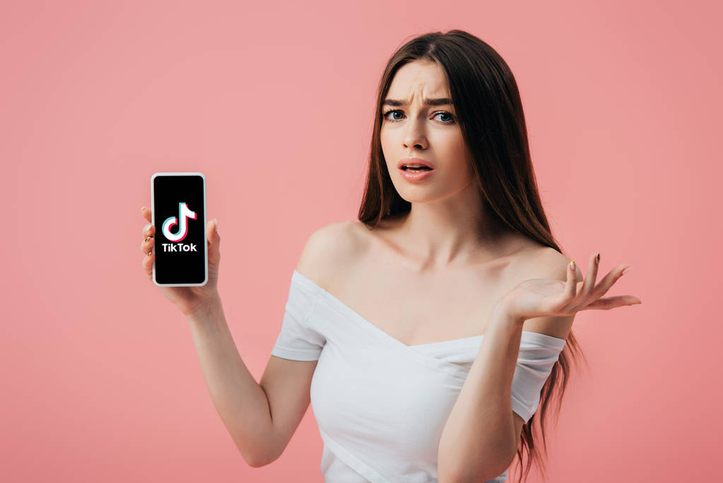 Kijów, Ukraina-6 czerwca 2019: piękne mylić dziewczyna trzyma smartphone z TIK tok aplikacji i pokazując wzruszyć gest izolowane na różowym - Zdjęcie, obraz