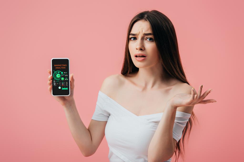 beautiful confused girl holding smartphone with online marketing analysis and showing shrug gesture isolated on pink - Photo, Image