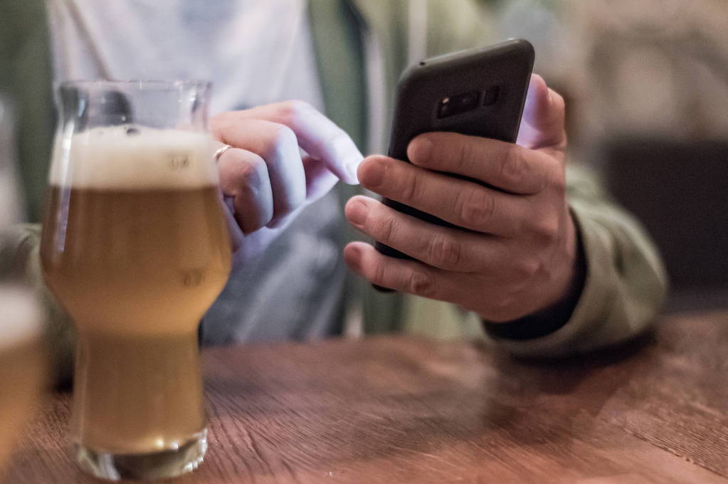 крупним планом рука людини тримає смартфон, п'є пиво і читає повідомлення в барі або пабі
 - Фото, зображення