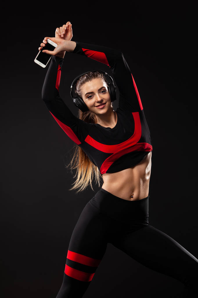 Beautiful young and fit woman in sportswear and headphones listening music from phone and smiling. Isolated on a black background. - Photo, Image