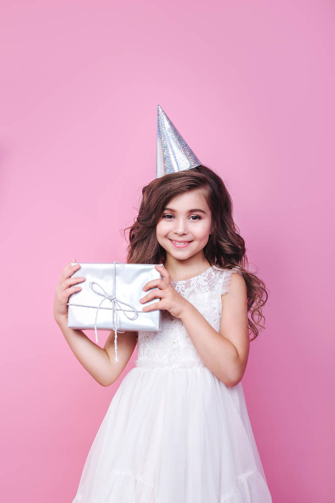 Jolie petite fille en robe et chapeau d'anniversaire regardant à l'appareil photo avec boîte cadeau dans les mains sur fond rose
 - Photo, image
