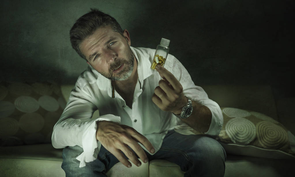 δραματικό πορτρέτο των νεαρών ελκυστική κατάθλιψη και σπατάλη χάπια εθισμένος άνθρωπος κρατώντας αντικαταθλιπτικά δισκία μπουκάλι κάθεται στον καναπέ στην έννοια του εθισμού στα ναρκωτικά - Φωτογραφία, εικόνα