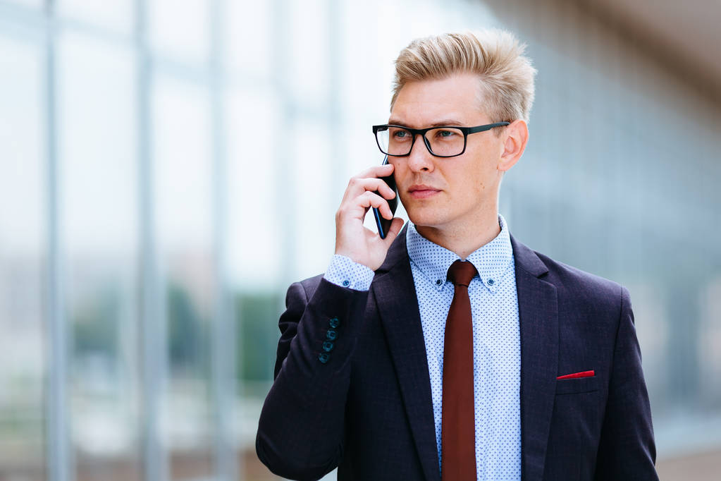 オフィスビルの前で携帯電話で話すメガネをかけた若いハンサムな白人ビジネスマン。屋外でスマートフォンを使用してトレンディな服を着た男性エグゼクティブマネージャー。現代技術の概念 - 写真・画像