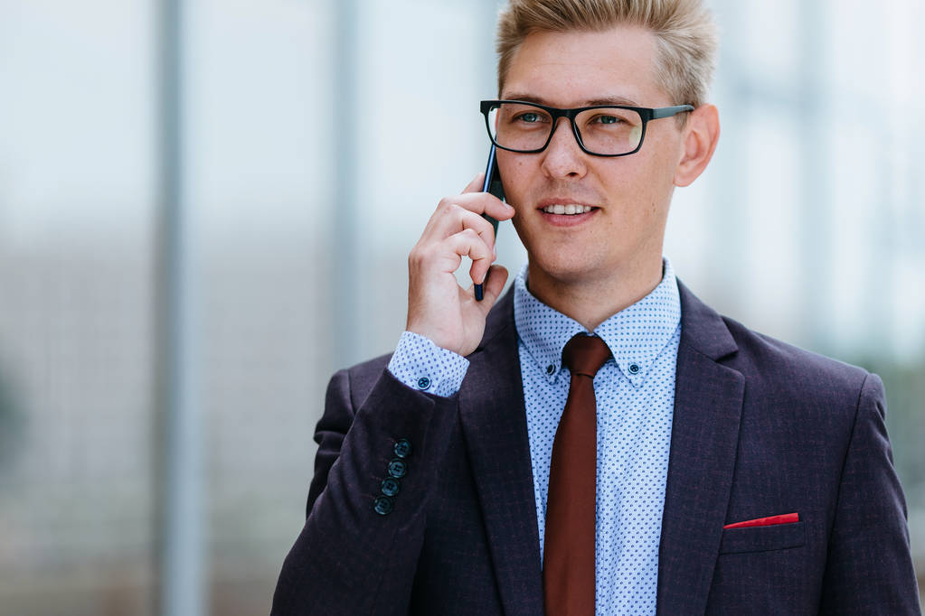 Jeune bel homme d'affaires caucasien en lunettes parlant sur un téléphone portable devant un immeuble de bureaux. Directeur exécutif masculin dans des vêtements à la mode en utilisant smartphone à l'extérieur. Concept de technologie moderne
 - Photo, image