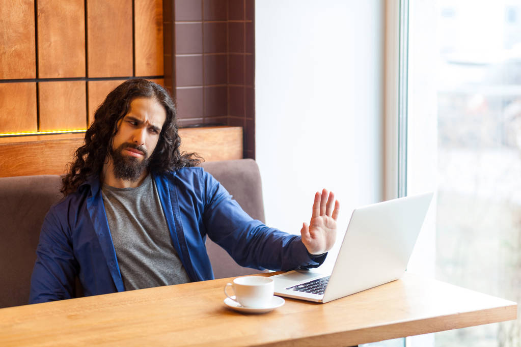 Serious handsome young adult man freelancer in casual style sitting in cafe and talking with opponent in laptop while showing stop hand gesture - Photo, Image